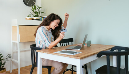 Cheerful smiling woman holding her fist up and credit card while sitting at table and looking at her laptop computer at home