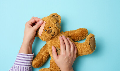 female hand holds a brown teddy bear and glues a medical adhesive plaster on a blue background,...