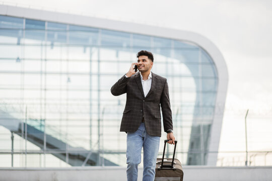 Portrait of young handsome Indian business man walking outside to station or airport talking on the phone and carrying suitcase.