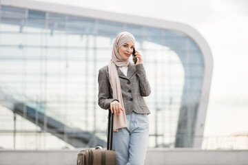 Beautiful young excited woman in hijab talking on the phone with friend, in anticipation of a wonderful trip and vacation. Arab lady standing with suitcase near airport