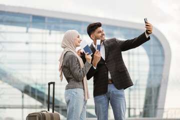 Joyful Muslim couple ready for trip, taking selfie outdoors near airport terminal, happy smile and gesturing. Young man and woman in hijab travel together, waiting for departure and maing video call