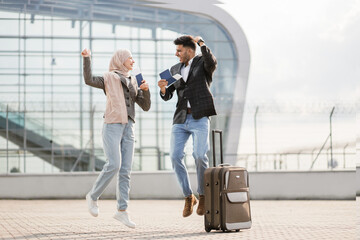 Happy excited couple, Muslim lady in hijab and handsome Arab man, are glad and jumping together, standing outside modern airport terminal and holding passports and tickets. Friends travel concept