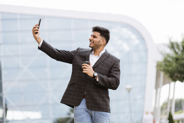 Cheerful good-looking Arabian businessman standing outdoors at new modern building, looking at mobile phone, making different emotions and gestures for selfie photo