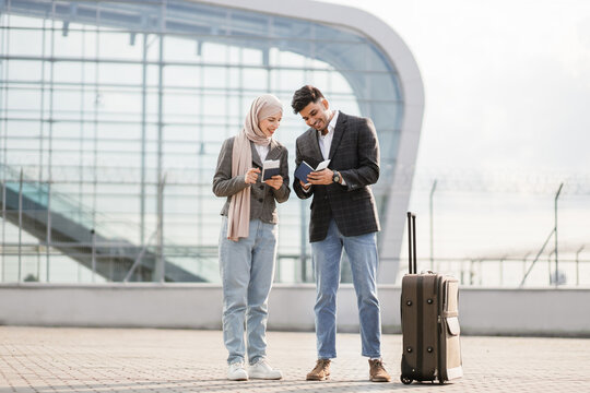 Business, trip and people concept. Happy business couple, Muslim woman in hijab, and Arab man, carrying suitcase outside airport, holding passports and tickets. Woman talking on phone