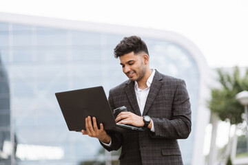 Fototapeta premium Focused young successful Arab businessman in formal wear, standing outside modern building and surfing internet on laptop. Confident bearded man enjoying fresh air while working outdoors.
