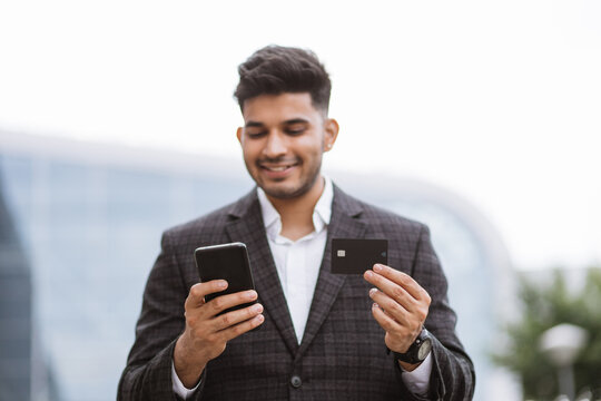 Business Trip Concept. Handsome Young Indian Man In Eyeglasses, Using Smart Phone And Holding Credit Card With Shopping Online. Male Freelancer Receives Payment Online