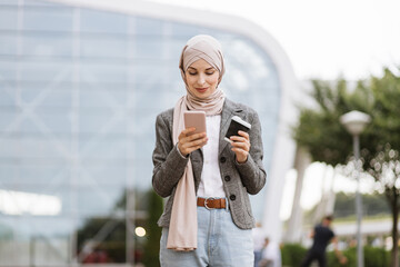 Pretty happy Muslim lady in hijab smiling at camera, standing outdoors with coffee to go and phone in hands. Likable Arab woman in headscarf walking on the street with take away coffee and phone