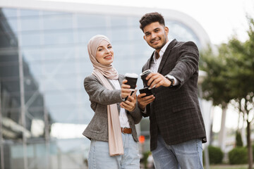 Smiling business couple, handsome Arab man and Muslim woman in hijab, standing outdoors in the street on coffee break, using phones and drinking take away coffee