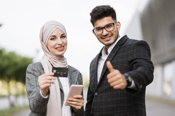 Smiling satisfied multiracial business couple buying online with credit card standing outside airport terminal and showing thumb up.