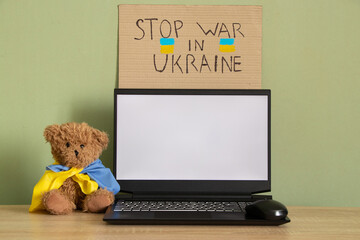 Teddy bear with flag of Ukraine near laptop with white screen on table at home, war in Ukraine, patriot, template, peace in Ukraine, laptop