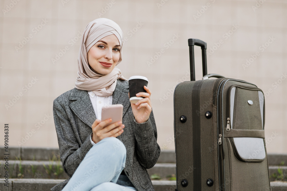 Wall mural young muslim woman in hijab dressed in jeans and jacket using phone and drinking take away coffee, w