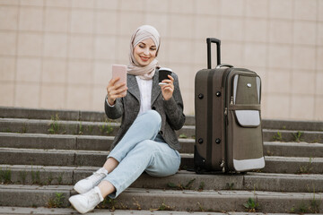 Focused young Muslim businesslady in hijab sitting on stairs and working on wireless laptop, while...