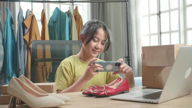 Asian Young Girl Online Seller Taking Photos With Mobile Phone While Using Computer For Selling Clothes At Home
