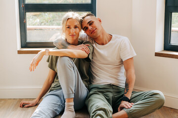 Young attractive lovers girl and guy sitting together on the floor look  at the camera.