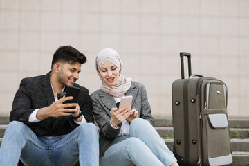 Cheerful smiling diverse business man and woman in hijab sitting on the stairs outdoors with smarphones and coffee, waiting for their business trip. Big travel bag on the stairs.