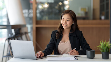 Beautiful young Asian businesswoman is smiling at her desk and taking notes with computer laptop on her desk, enjoying work.