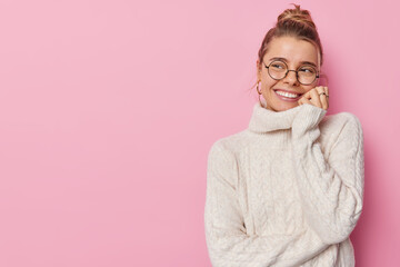 Horizontal shot of pretty young woman with hair bun smiles gently looks away with glad expression listens something pleasant wears spectacles and knitted sweater poses indoor against pink wall