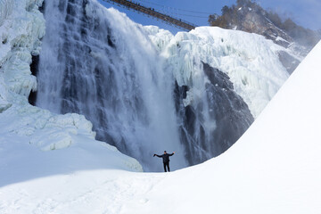 Low angle side view of middle-aged man standing dressed in black with arms raised on ice formation at the foot of the Montmorency Fall during a beautiful sunny winter day, Quebec City, Quebec, Canada
