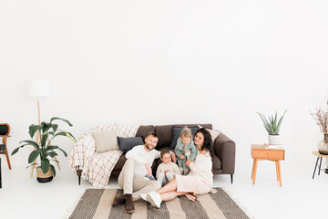 Obraz premium Stylish happy parents sitting near the sofa with joyful little kids, smiling, spending weekends at home. Charming father with pretty mother playing with cute daughter and son, family concept