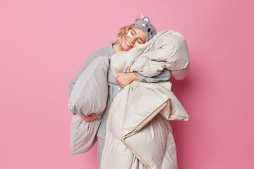 Relaxed young European woman sees sweet dreams embraces soft duvet leans on pillow applies beauty patches under eyes enjoys healthy sleep isolated over pink background. Cozy morning concept.