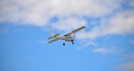 Pequena avioneta branca a levantar voo, vista por baixo de e trás, céu azul com nuvens brancas - obrazy, fototapety, plakaty