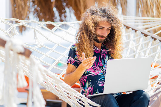 long curly haired man sitting on the hammock near the beach and working laptop vacation