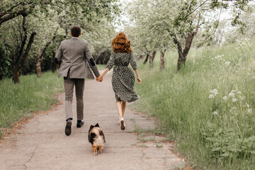 a couple in retro style running along a garden path with a small dog behind them