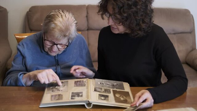 Elderly woman looks at souvenir photo album with her daughter. Senior woman sitting in the livingroom looking at photographs with daughter. Brain training. Short term memory enhancing activities.
