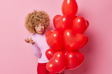 Indignant frustrated curly woman shrugs shoulders looks puzzled at camera holds big bunch of heart balloons doesnt like something purses lips has leaked makeup isolated over pink background.