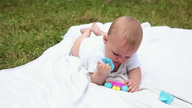 Little newborn girl lying on tummy chewing rattle on blanket, summer day outdoor. Infant having fun outdoors. Infant baby child resting playing learning to crawl. Motherhood teething, babyhood concept