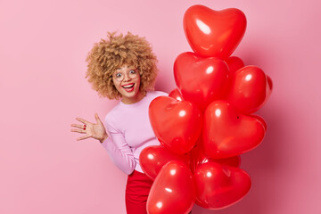 Happy curly haired woman looks from bunch of heart balloons keeps palm raised feels joyful wears spectacles and casual clothes poses against pink background glad to celebrate Valentines Day.