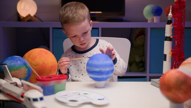 Blond boy paint planet solar system Neptune with colorful paint sitting home table in evening, planet solar system, spaceships and space shuttle from constructor around. Cosmonautics Day on April 12.