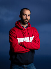 Portrait of bearded caucasian young man dressed in red and blue sweatshirt and jeans looking at camera at blue hour with flash