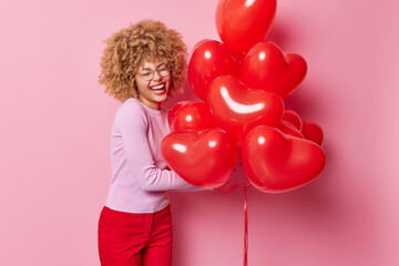 Indoor shot of happy young woman with curly hair laughs out gladfully enjoys celebration holds bunch of red heart balloons feels very happy receives congratulations on birthday or Valentines Day