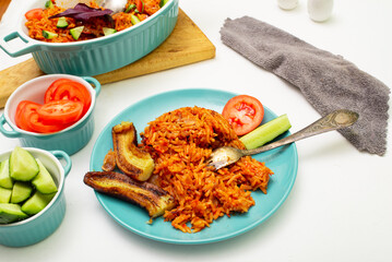 Jollof rice with fried banana on a plate. Rice with tomatoes, onions, spices. Fresh vegetables. White background.