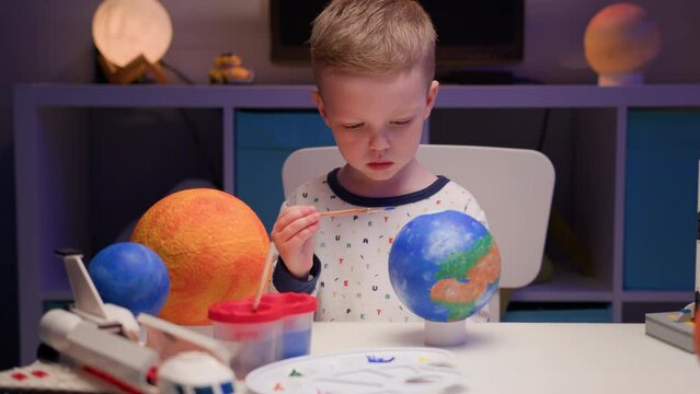 Blond boy paint planet solar system Earth with colorful paint sitting home table in evening, planet solar system, spaceships and space shuttle from constructor around. Cosmonautics Day on April 12.