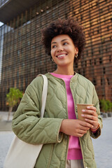Outdoor shot of curly haired woman wears jacket carries bag holds paper cup of coffee enjoys...