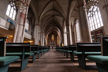 Das historische Kirchenschiff einer alten Kirche in Marburg mit hohen Säulen aus Sandstein und von der hohen Decke hängen riesige Kronleuchter, im Kirchgang stehen Sitzbänke mit grünem Samt bezogen
