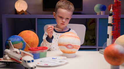 Blond boy paint planet solar system Jupiter with colorful paint sitting home table in evening,...