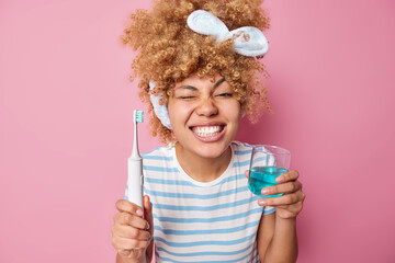 Dental health concept. Positive woman with combed curly hair holds electric brush and glass of mouthwash brushes her perfect teeth winks eye wears casual striped t shirt isolated on pink wall