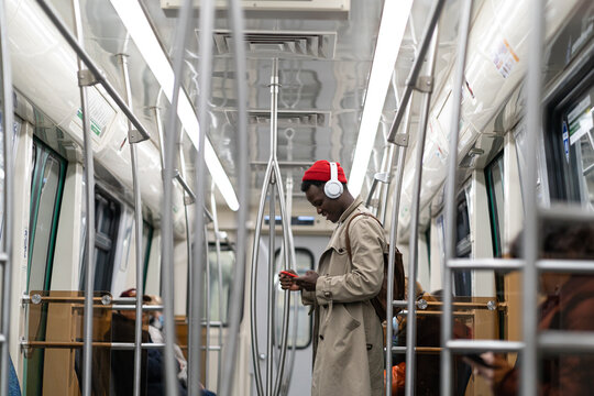 Young african american hipster man wear headphones, listening music, using smartphone in subway train. Black student male using mobile phone, chatting in social media in public transportation. 