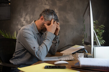 un homme de 50 ans est en télétravail chez lui dans son bureau devant son ordinateur. Il travaille beaucoup et il est fatigué. Il se prend la tête dans les mains.