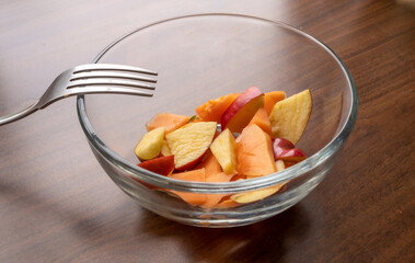 Tazón de vidrio sobre mesa de madera con poca fruta picada y tenedor a la luz de la ventana sobre mesa de madera 