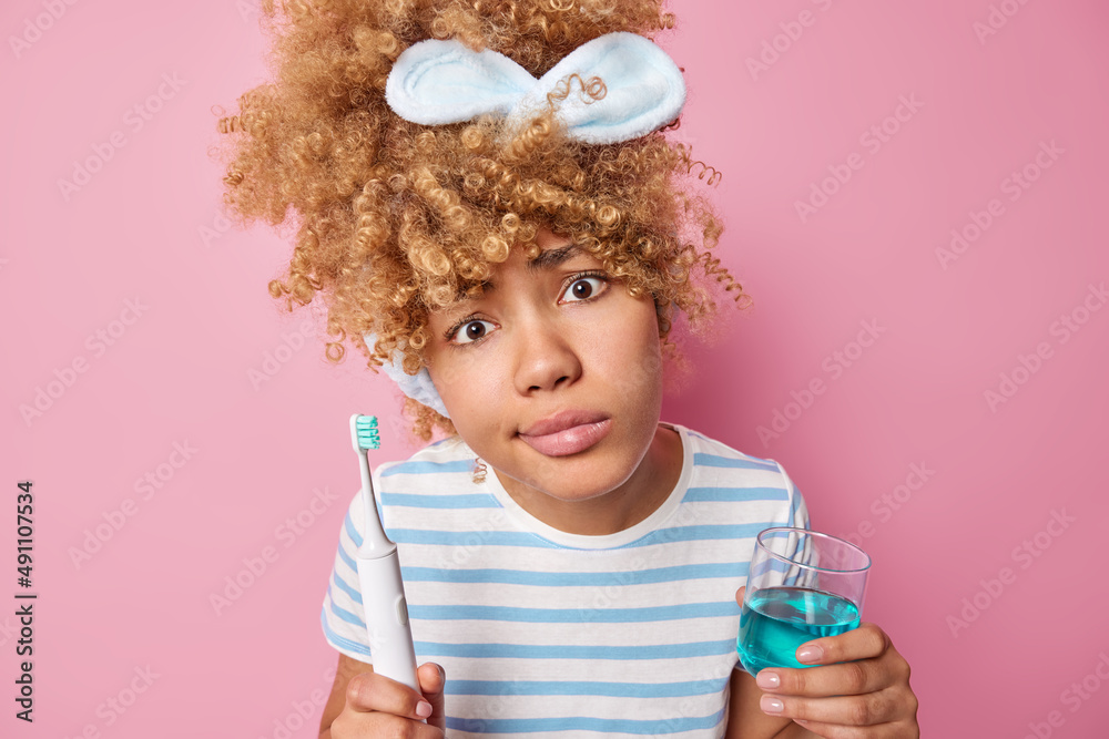 Wall mural Indignant curly haired woman holds glass of mouthwash and toothbrush for fresh breath wears headband and casual striped t shirt poses against pink background. Daily hygiene procedures concept