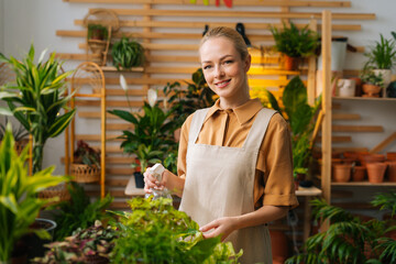 Portrait of smiling female florist in apron spraying water on houseplants in flower pots by sprayer, looking at camera. Happy young woman sprinkling green leaves of house flowers using spray bottle.