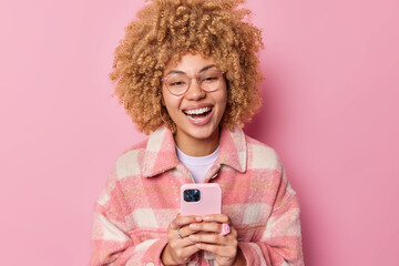 Indoor shot of happy curly haired woman laughs joyfully dressed in casual checkered jacket uses mobile phone connected to wifi types feedback enjoys cellphone messaging isolated over pink background