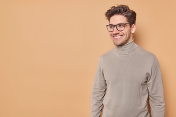 Studio shot of handsome European man smiles positively being in good mood focused somewhere wears spectacles and turtleneck poses against beige background with copy space for your promotion.