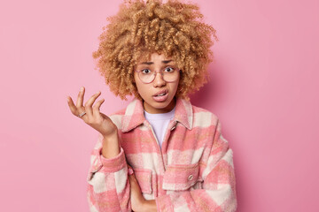 Indignant woman with curly hair raises hand and shrugs shoulders purses lips wears checkered jacket has hesitant expression isolated over pink background. So what should I do in this situation
