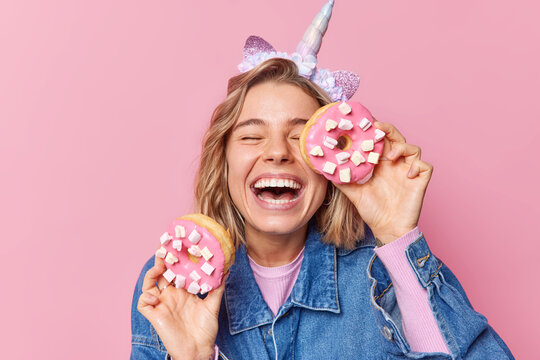 Overjoyed Fair Haired Woman Smiles Broadly Holds Delicious Doughnuts With Marshmallow Has Sweet Tooth Enjoys Eating Yummy Dessert Wears Unicorn Headband And Denim Jacket Isolated Over Pink Wall