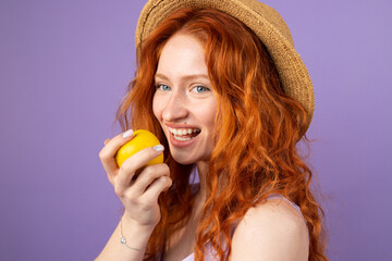 Young redhead woman with blue eyes wearing a straw hat smiling widely holding an apple ready to eat on purple background.